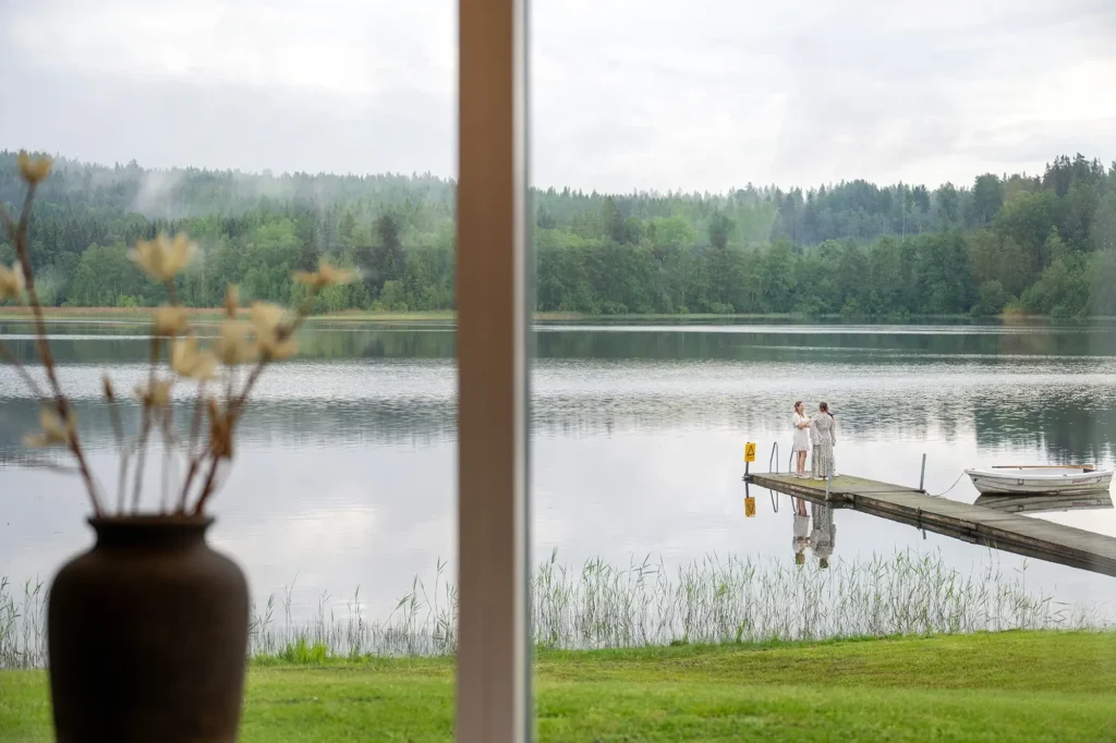 Utsikt genom boende fönstret mot sjö, brygga, båt och två personer. Närmare Viredaholmssjön är svårare att bo. Wiredaholm Golf & Konferens