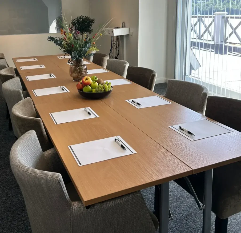 Konferensrum med långt mötesbord, stolar, skrivunderlägg, frukt och blommor.